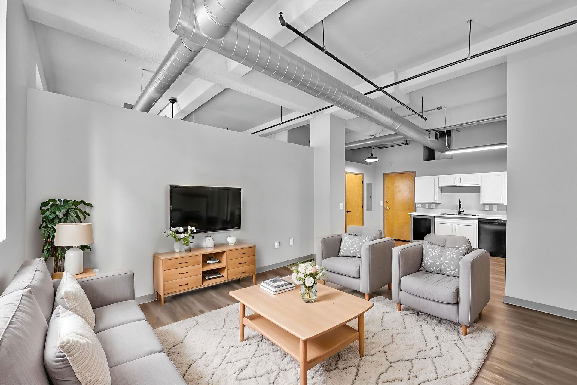 A bright living room with gray furniture, wood coffee table, TV console, and open kitchen in a modern, industrial space.