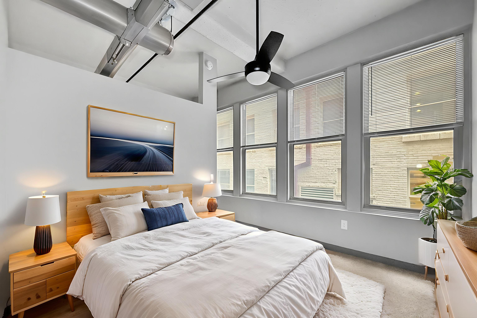 A modern bedroom featuring a bed with neutral linens, a wood headboard, framed artwork, and large windows with blinds.