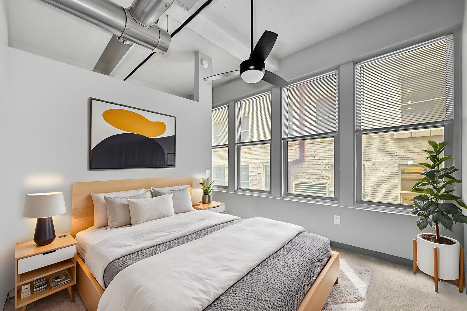 A modern bedroom with a queen bed, light wood nightstand, abstract art, ceiling fan, and large windows with blinds.