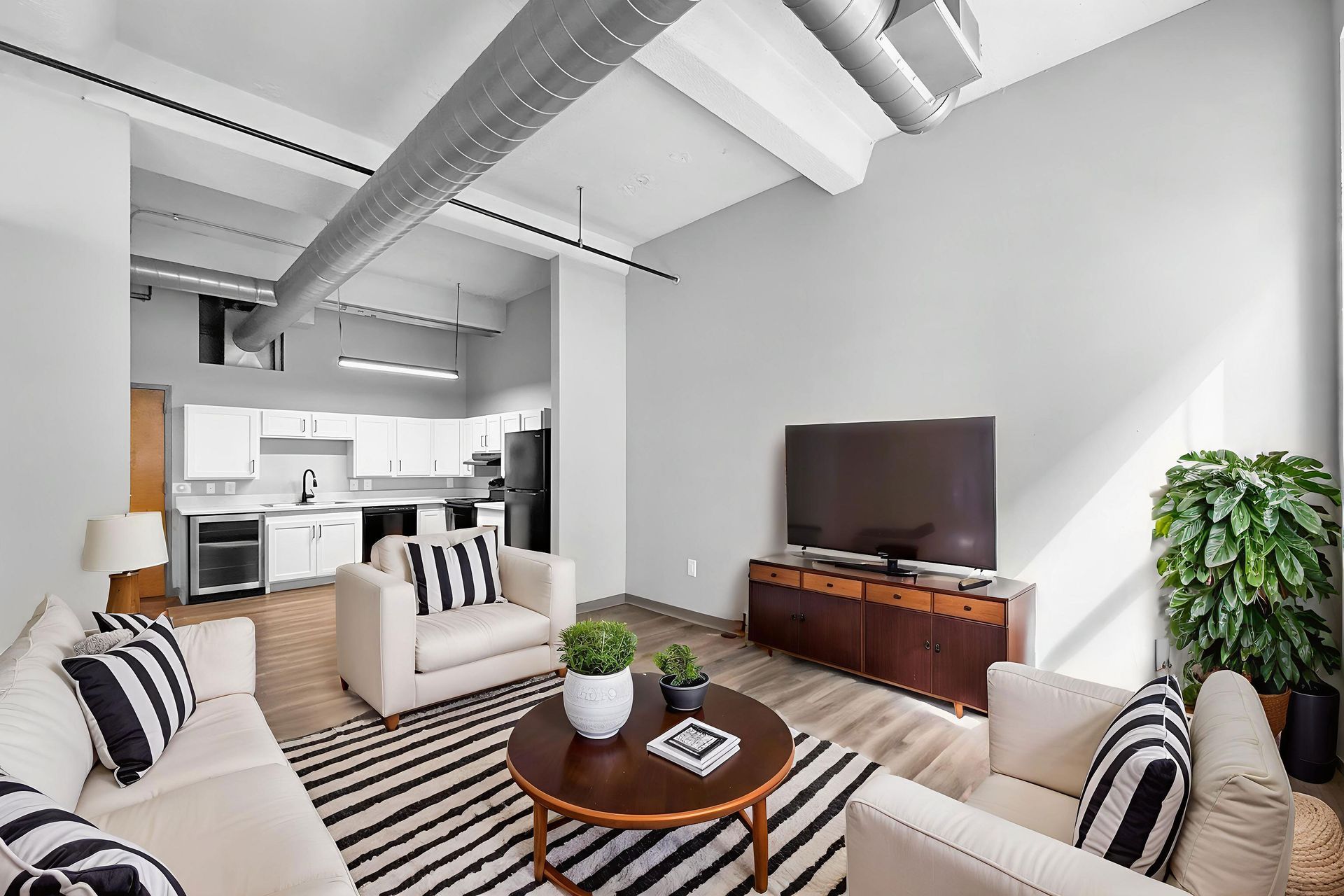 A modern open-plan living room with white furniture, black and white striped pillows, a wood coffee table, and a TV.