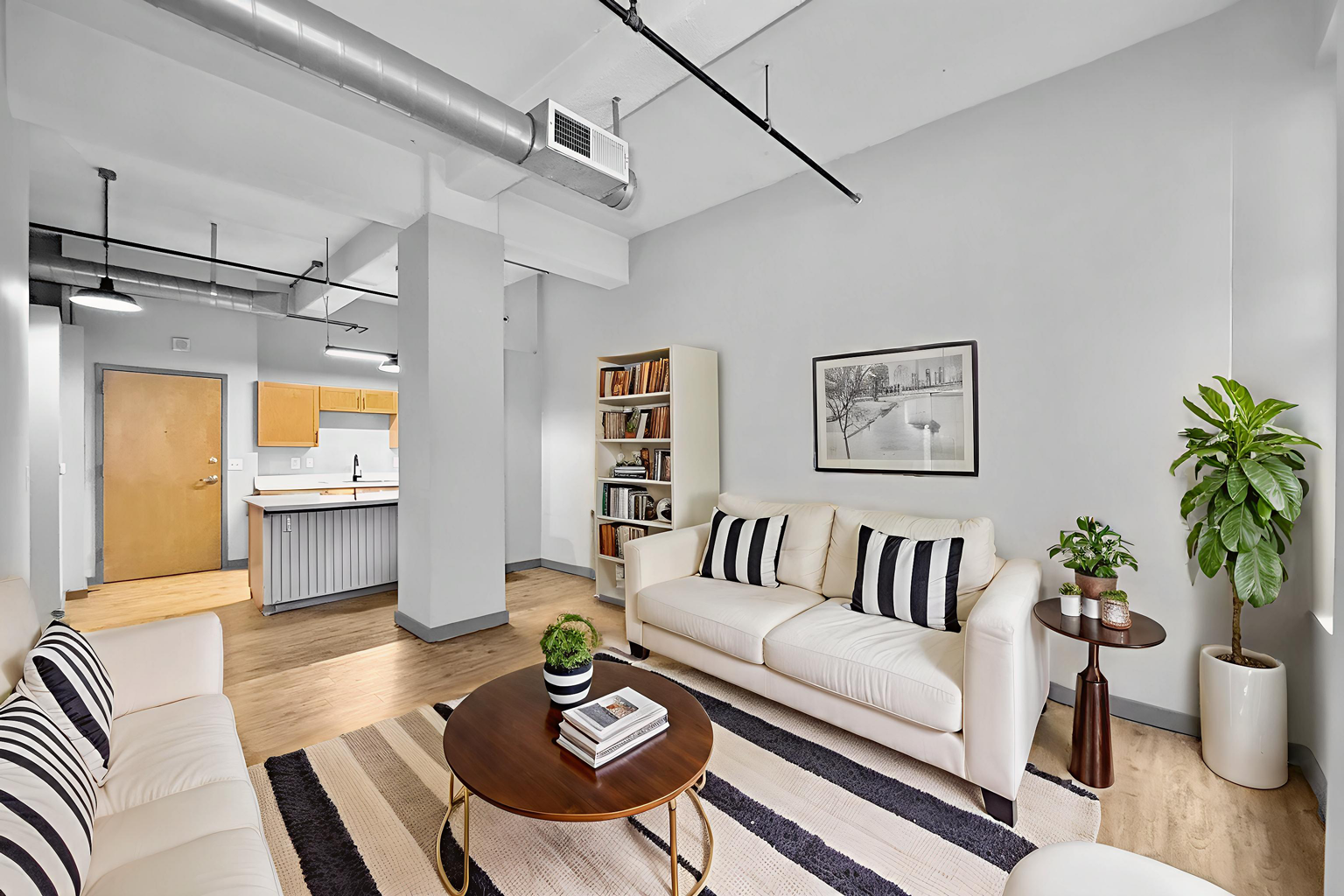 A modern living room with a beige sofa, round wood coffee table, striped area rug, white bookshelf, and a potted plant.
