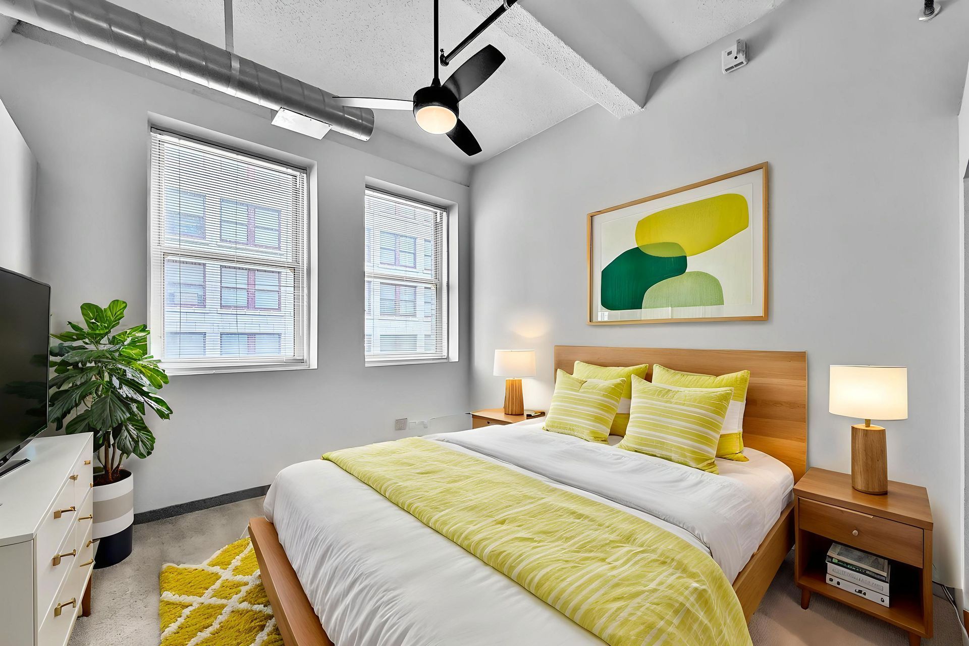 A bright modern bedroom with a queen bed, yellow accents, abstract art, twin windows, a ceiling fan, and wooden furniture.