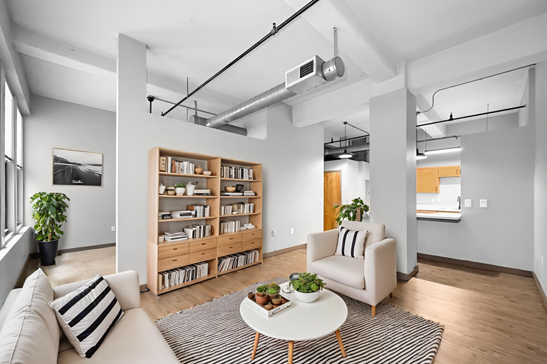 A bright, modern living room with light wood floors, a beige sofa, matching armchair, bookshelves, and a round table.