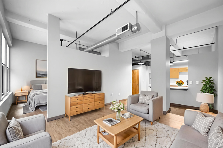 A bright, open-concept living room and bedroom featuring modern industrial decor, light wood furniture, and gray sofas.
