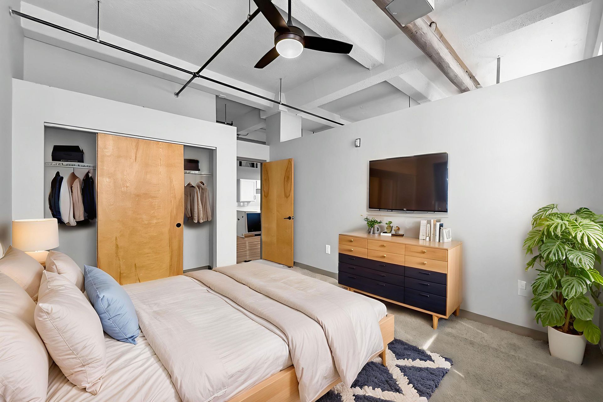Modern bedroom with concrete floors, a bed with light bedding, wooden closets, a wall-mounted TV, and a large plant.