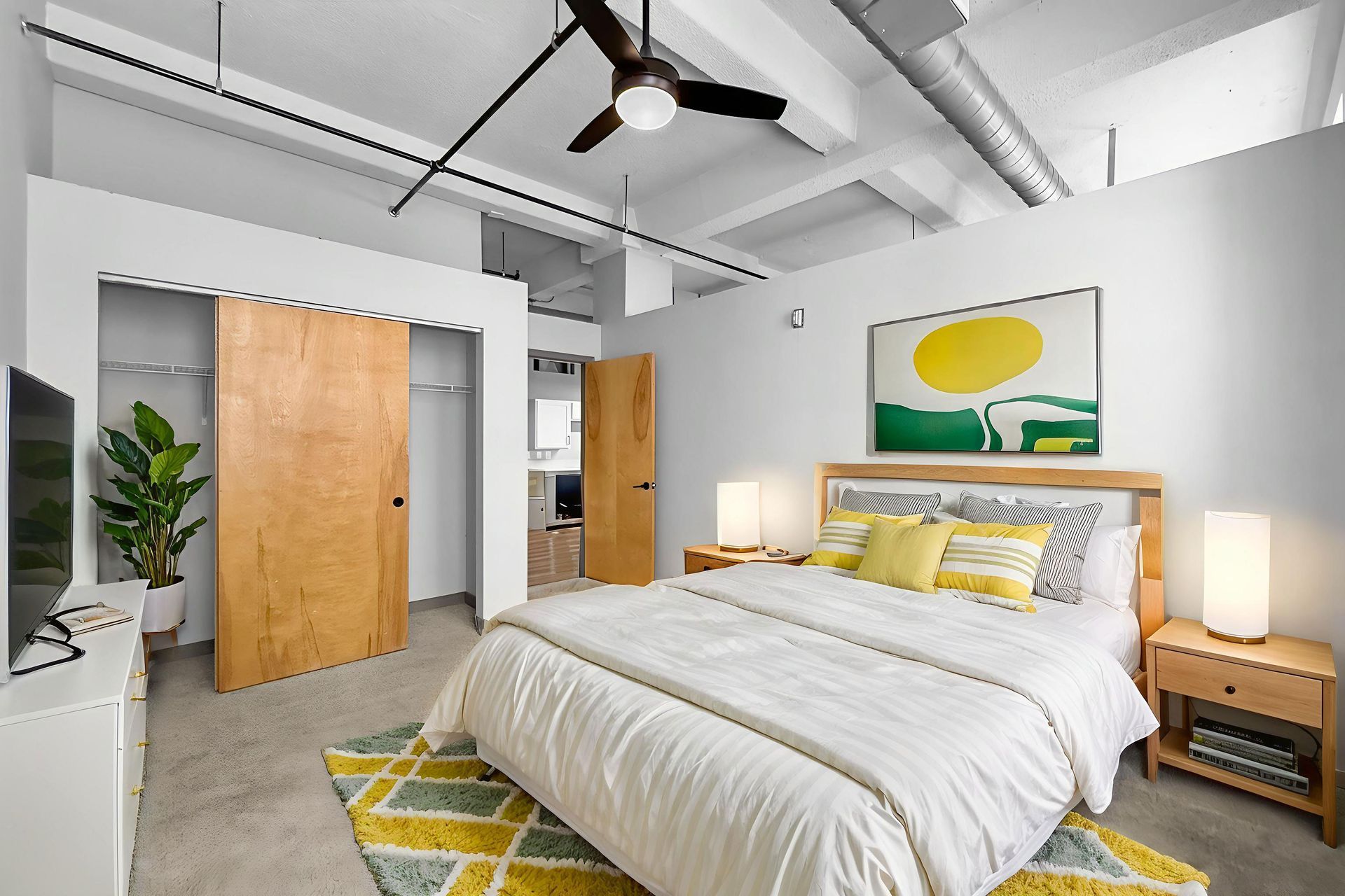 A modern, sunlit bedroom with a queen bed, wood closet doors, yellow-patterned rug, and ceiling fan in an industrial loft.