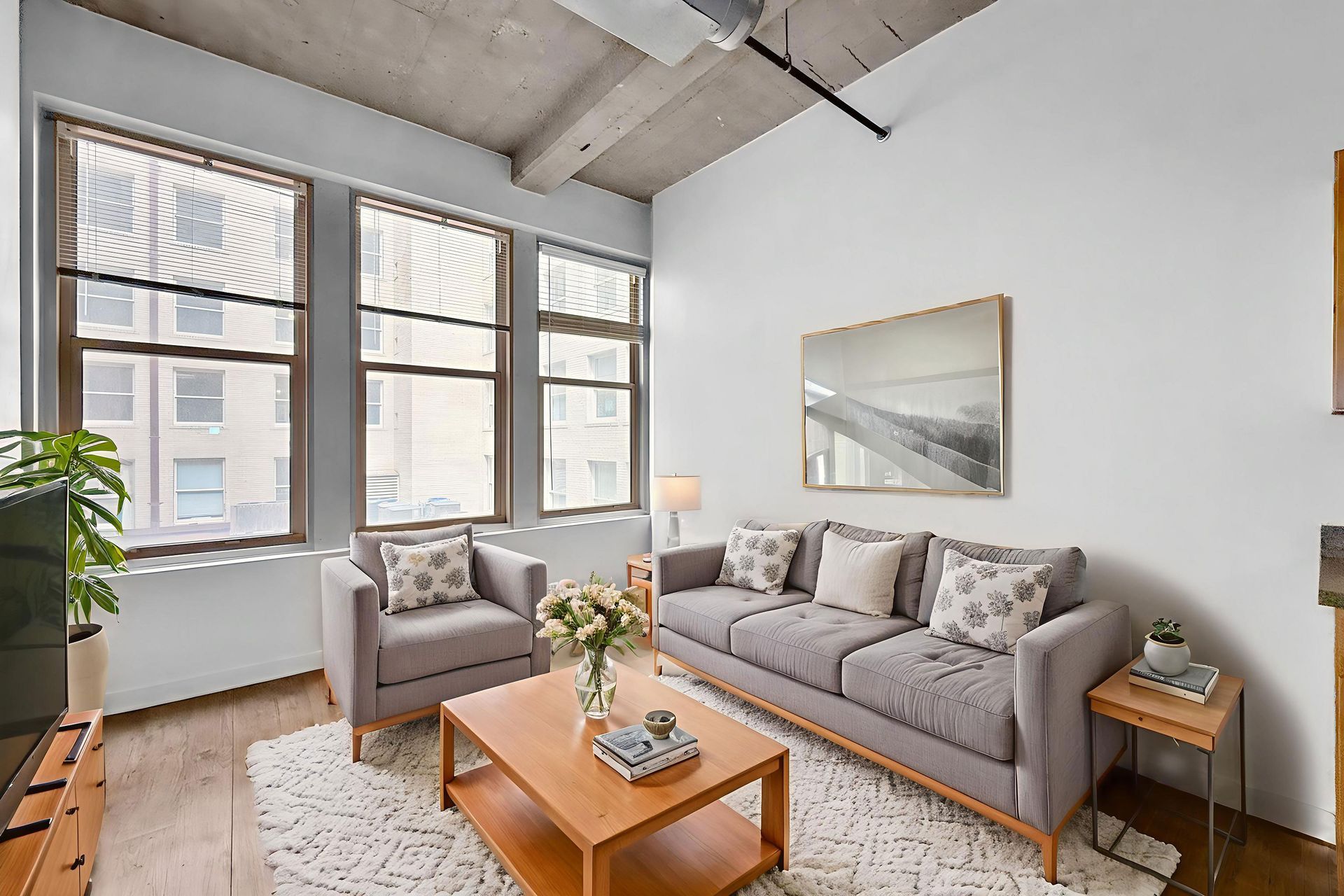 A modern living room with gray furniture, a wooden coffee table, large windows, and a high, unfinished concrete ceiling.