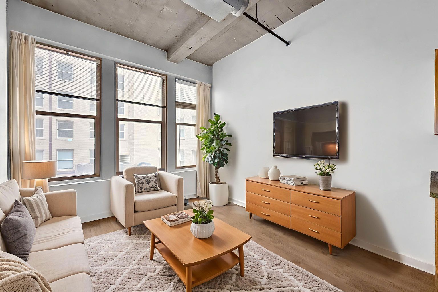A bright living room with a sofa, armchair, coffee table, wood dresser, TV, and large windows with cream curtains.