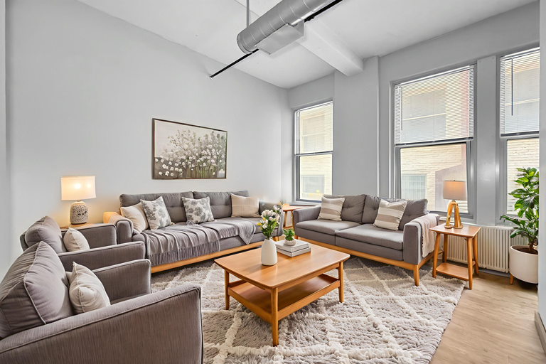A bright, minimalist living room with two gray sofas, matching armchairs, a wooden coffee table, and large windows.
