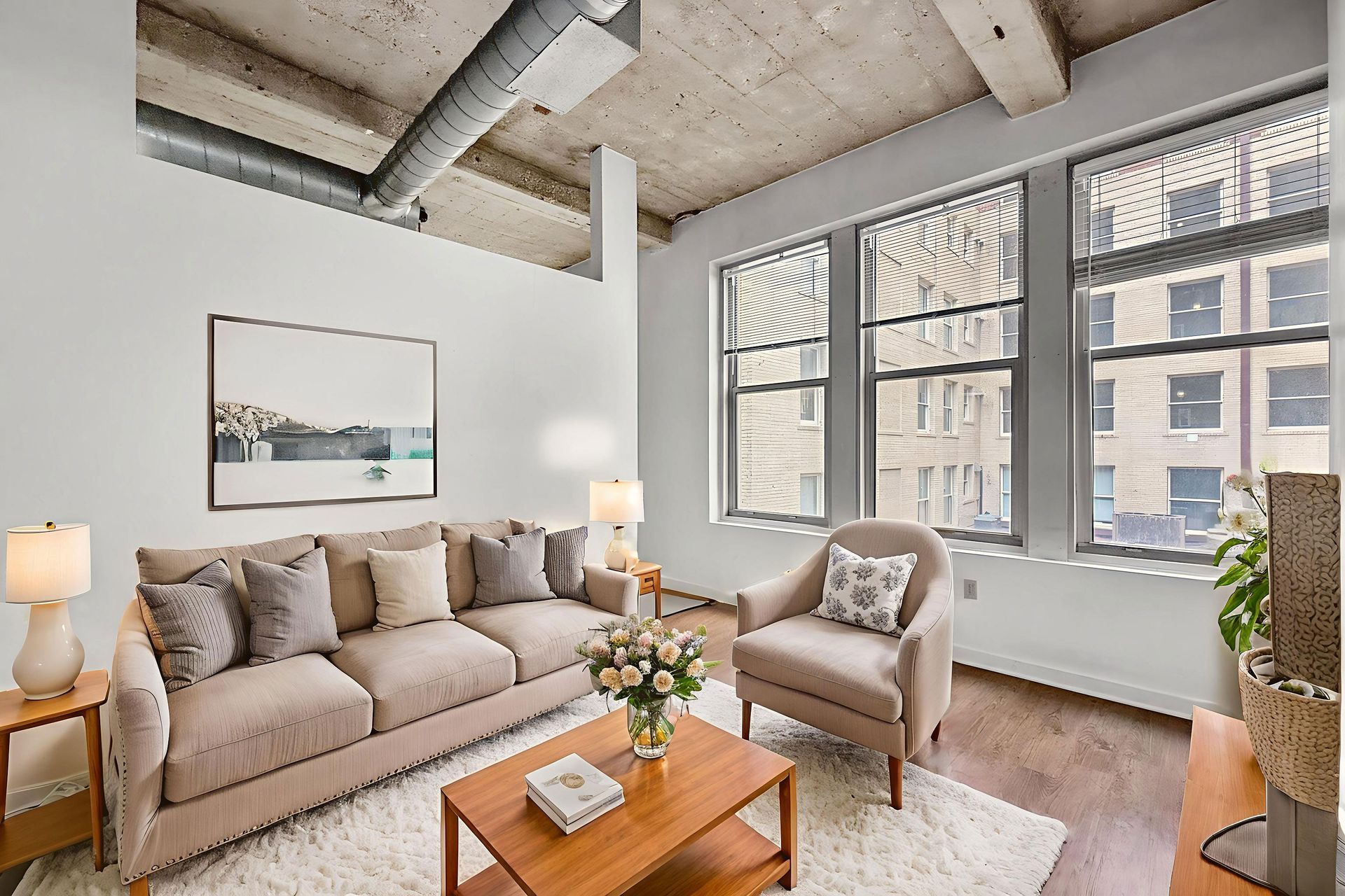 A beige sofa and armchair in a modern living room with large windows, a wooden coffee table, and a concrete ceiling.