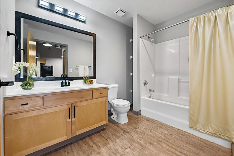A bathroom with a wooden vanity, a large framed mirror, a white toilet, and a bathtub with a beige curtain.