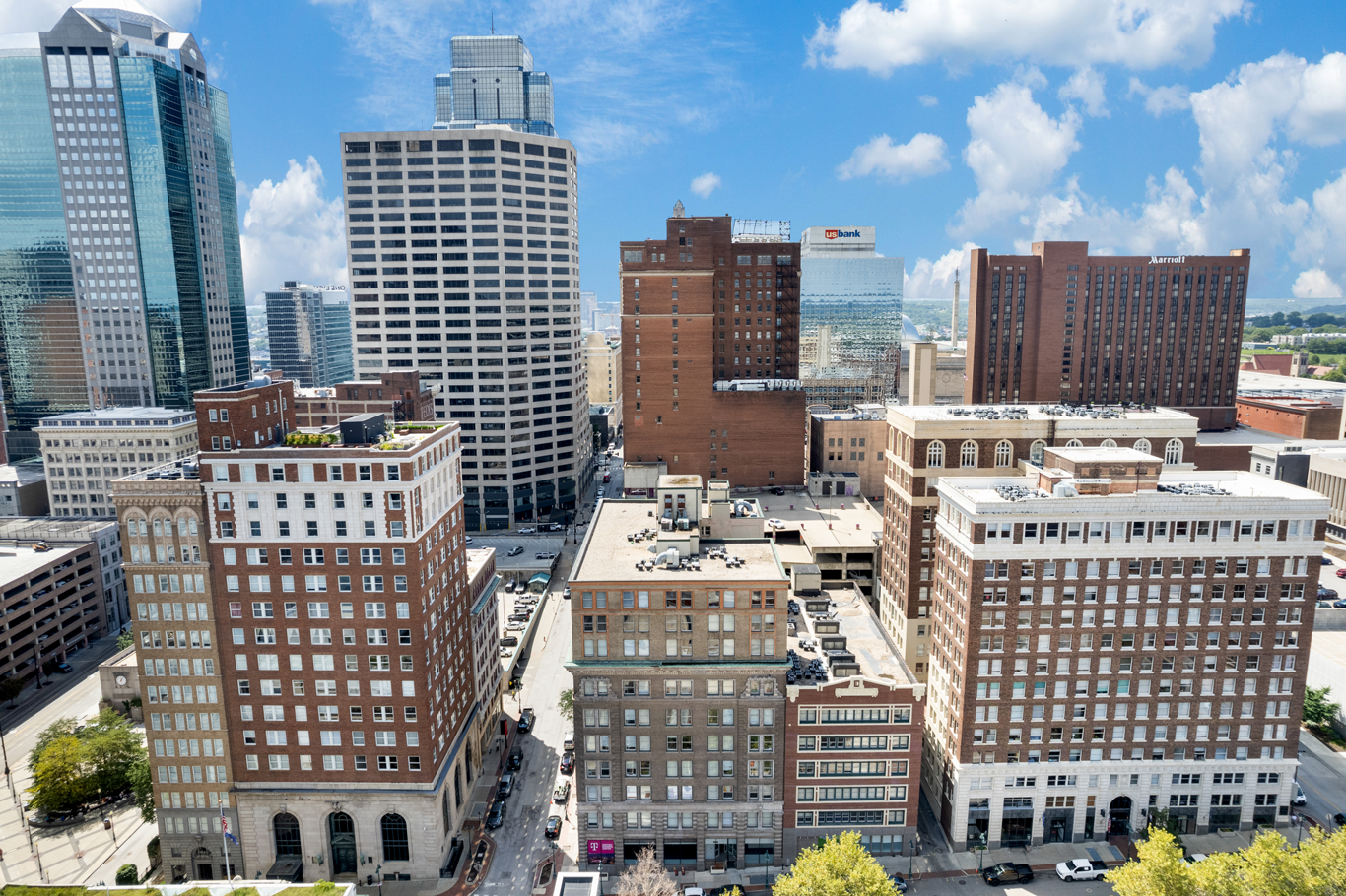 An aerial view of a city skyline with various brick and glass buildings under a bright blue sky with scattered clouds.