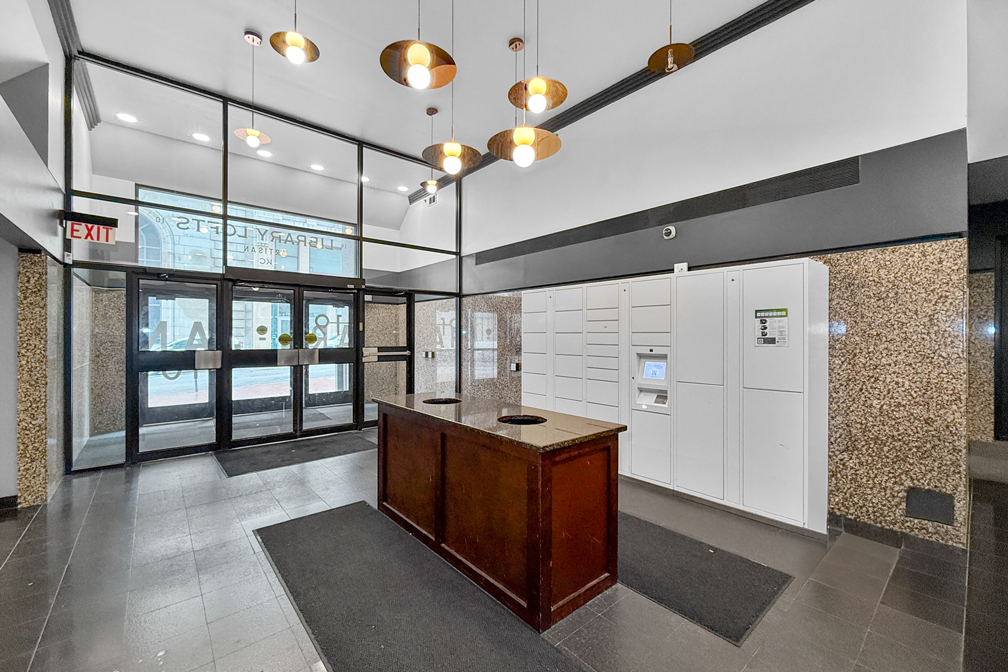 A bright building lobby with glass entrance doors, pendant lights, a dark wood reception desk, and white parcel lockers.