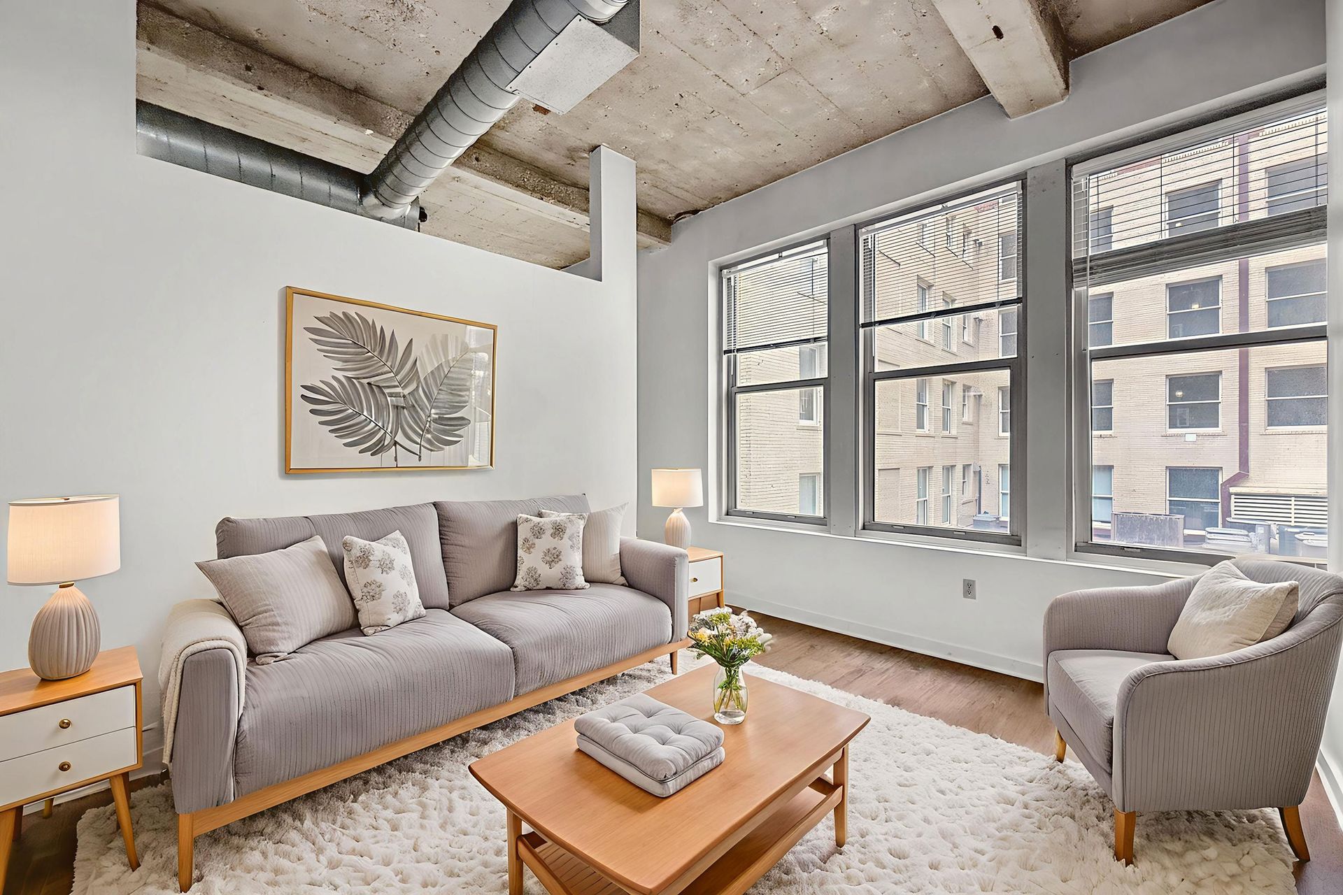 A bright living room with a grey sofa and armchair, a wooden coffee table, rug, and windows against a concrete ceiling.