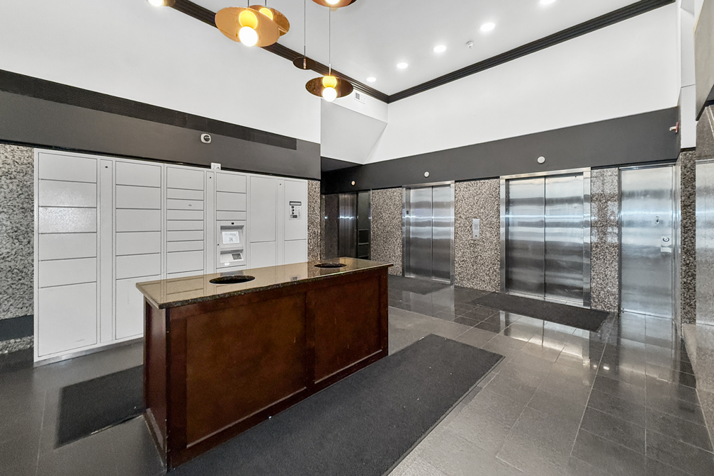 An interior lobby with a dark wood front desk, a wall of white parcel lockers, and two metal elevators with gray tiles.