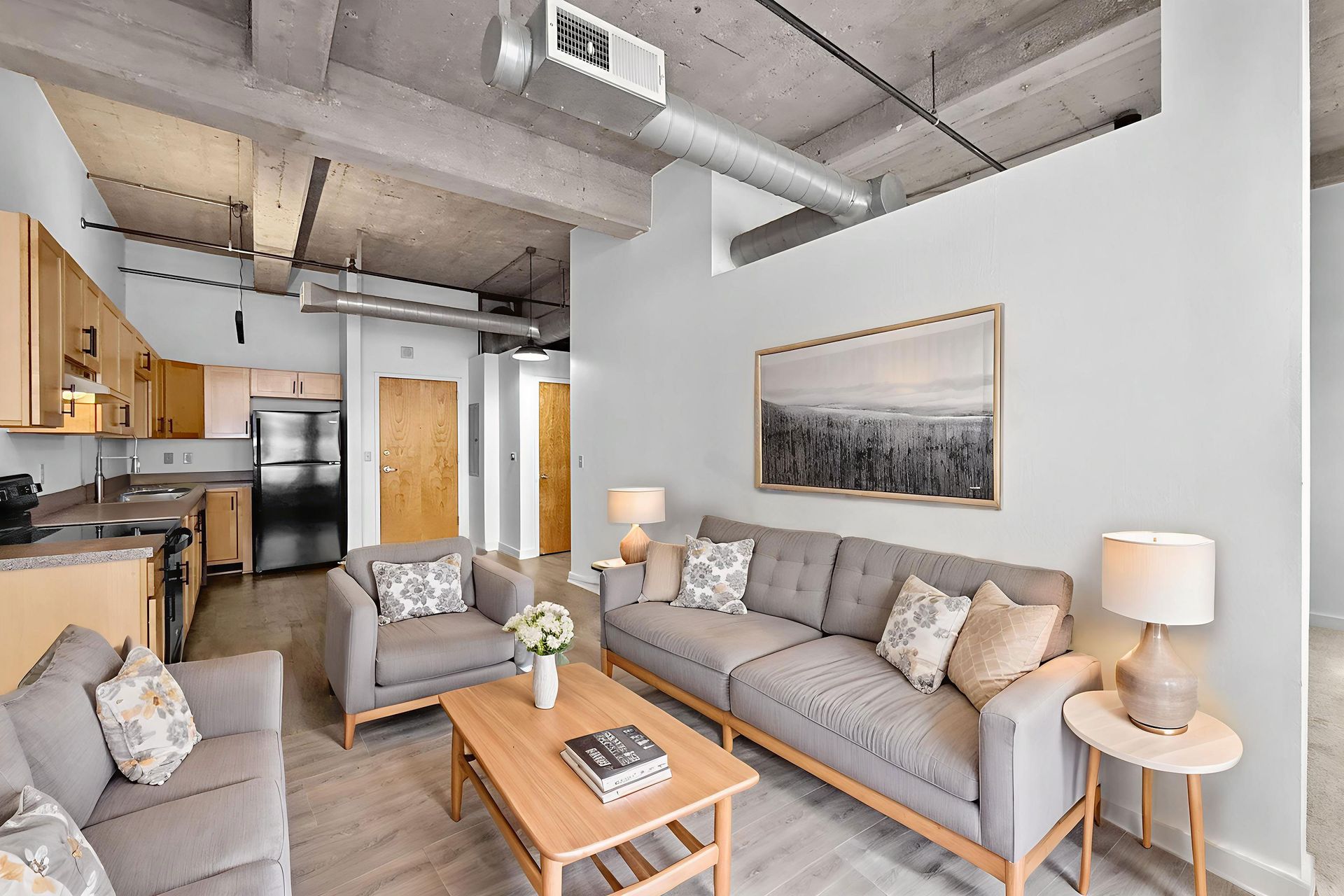 A modern, open-concept apartment living room with a grey sofa, armchair, coffee table, and a kitchen in the background.
