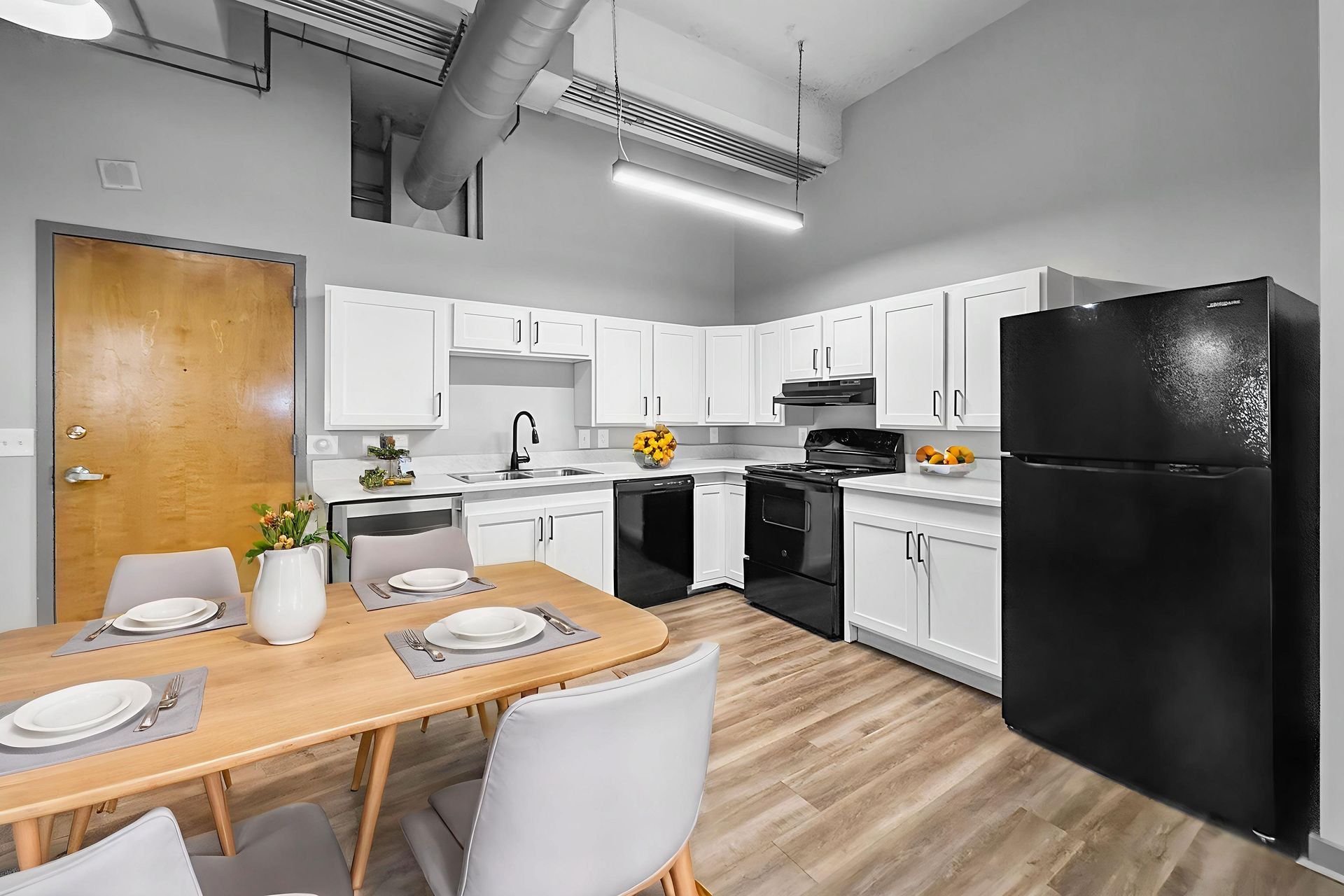 Modern kitchen with white cabinets, black appliances, a wooden dining table, and light wood floors in an open space.