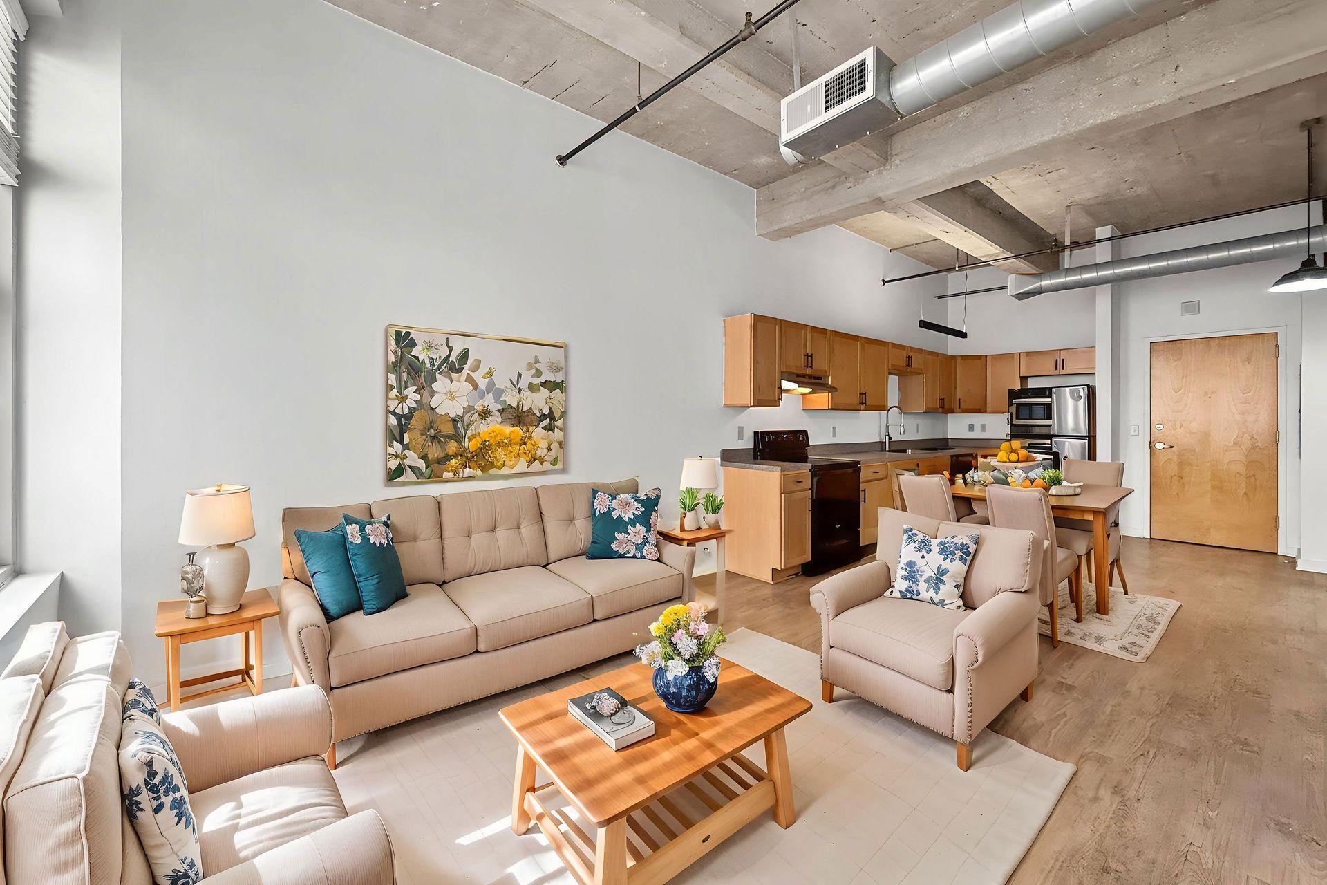 A bright, open-concept studio apartment featuring a beige couch, armchair, wooden coffee table, and kitchen in the back.