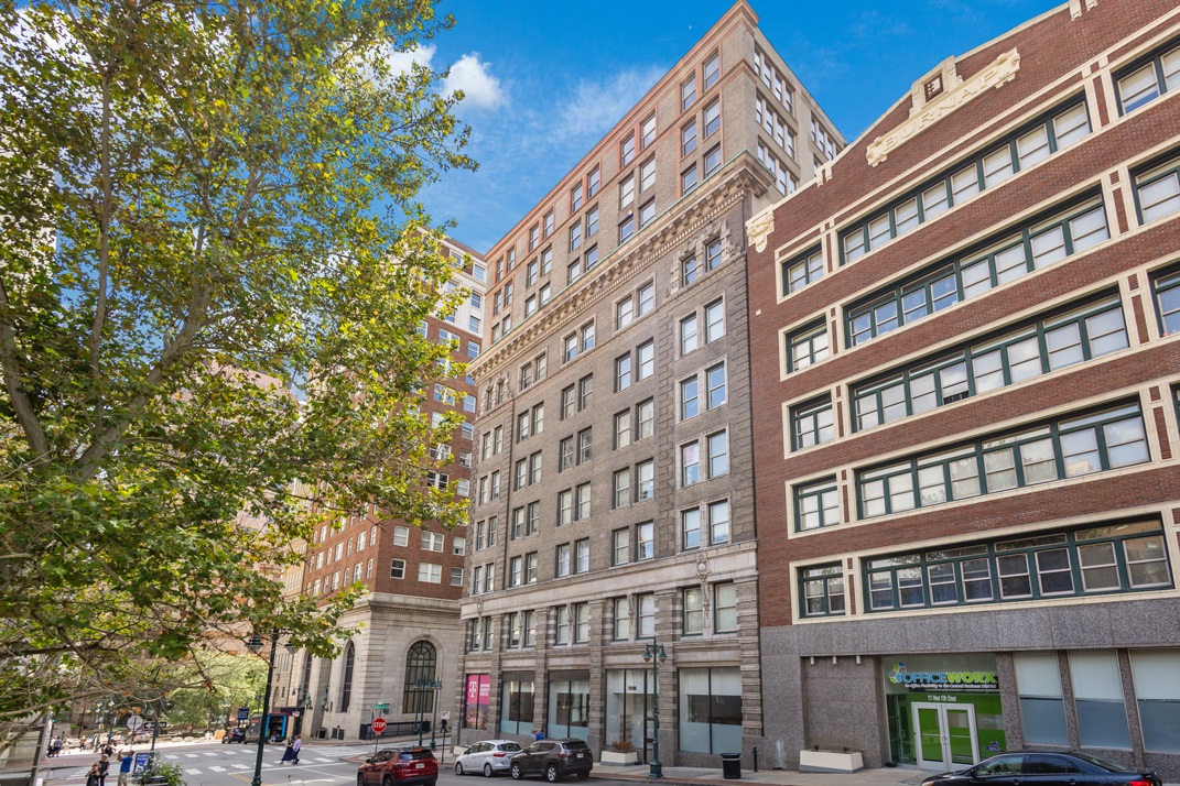 A low-angle view of two brick city buildings under a bright blue sky, with a lush green tree in the foreground.
