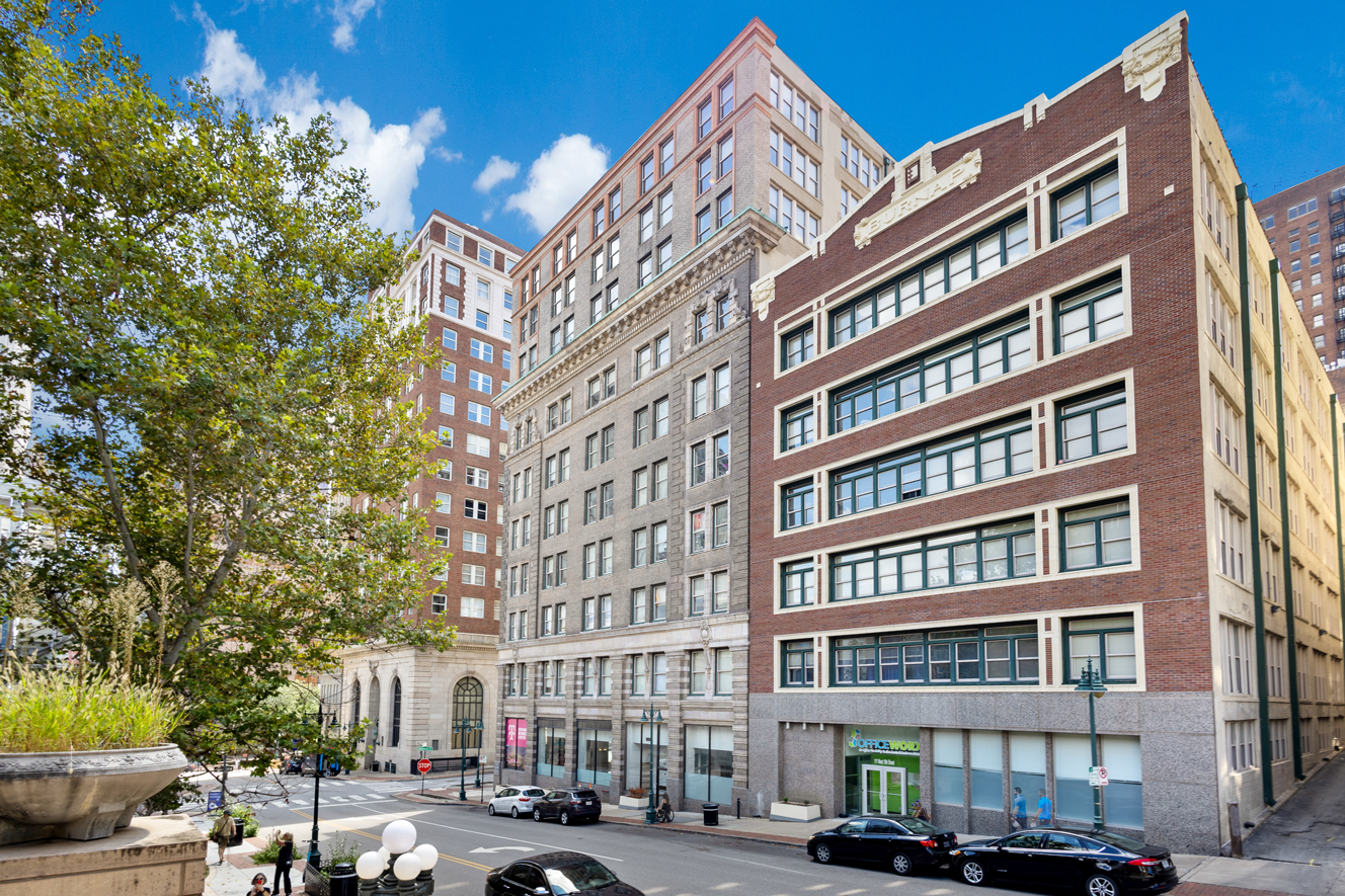 A street-level view of several historic brick and stone urban buildings on a sunny day with trees in the foreground.