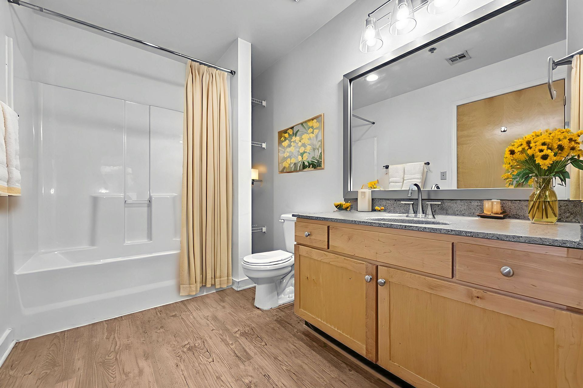 A bathroom with light wood cabinets, a bathtub with a beige curtain, a mirror, and a vase of sunflowers.