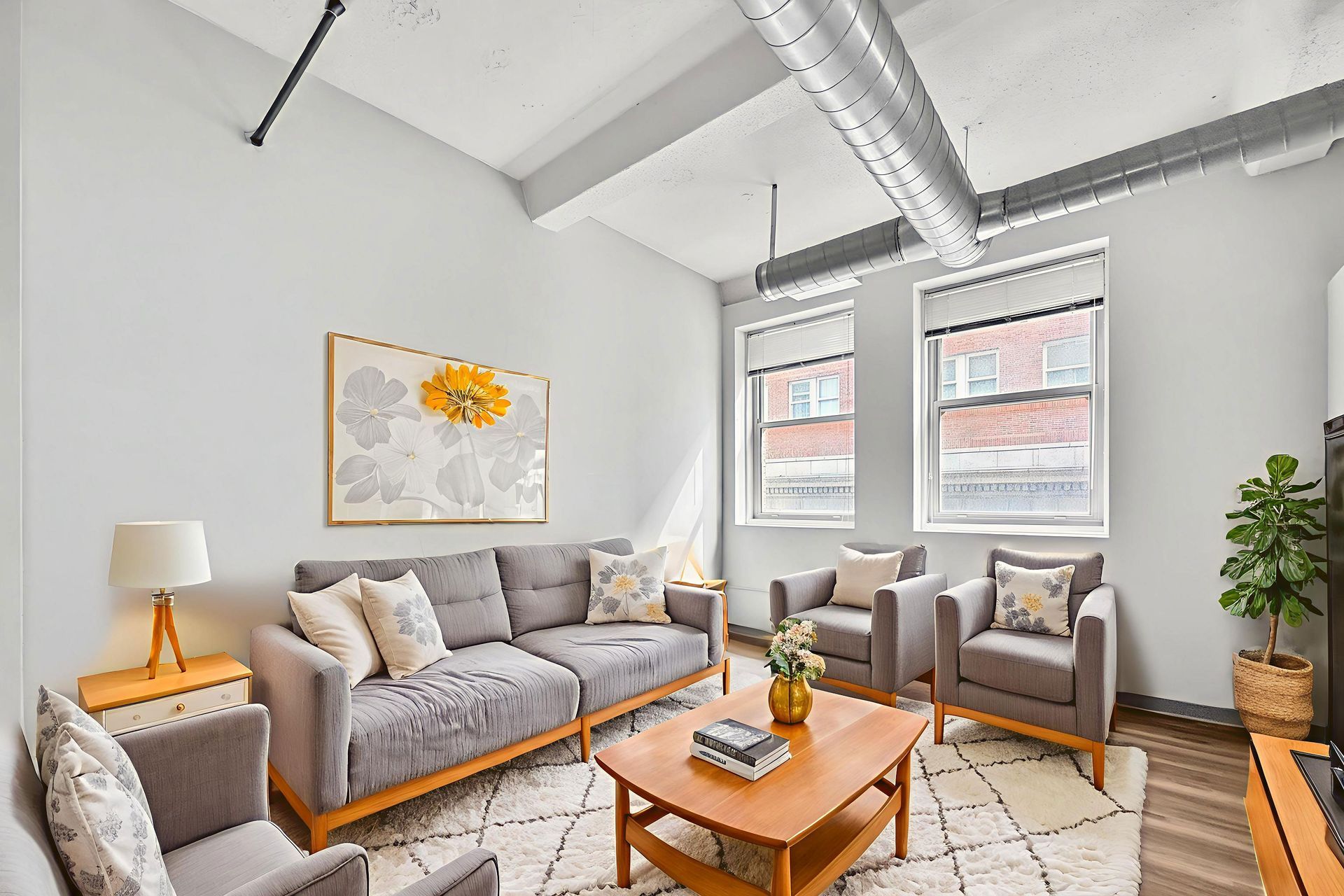 A bright, industrial-style living room with a grey sofa, matching armchairs, a wood coffee table, and large windows.