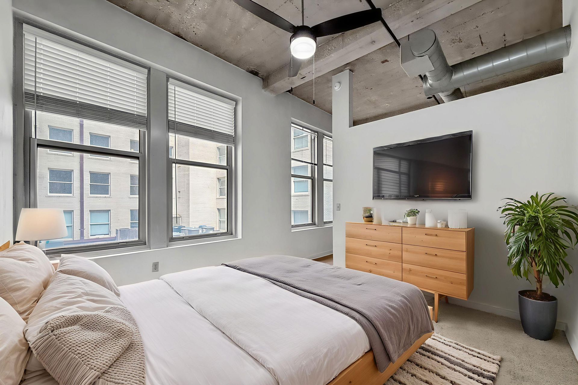 A modern bedroom with a bed, light wood dresser, wall-mounted TV, concrete ceiling, and large windows with blinds.