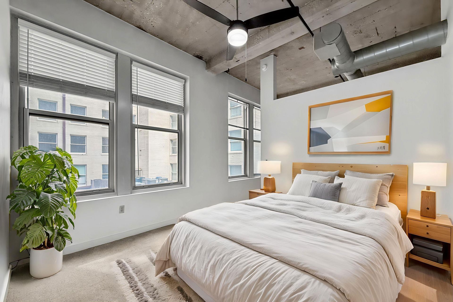 Modern bedroom with a white bed, beige carpeting, a large potted plant, and exposed concrete ceilings with large windows.