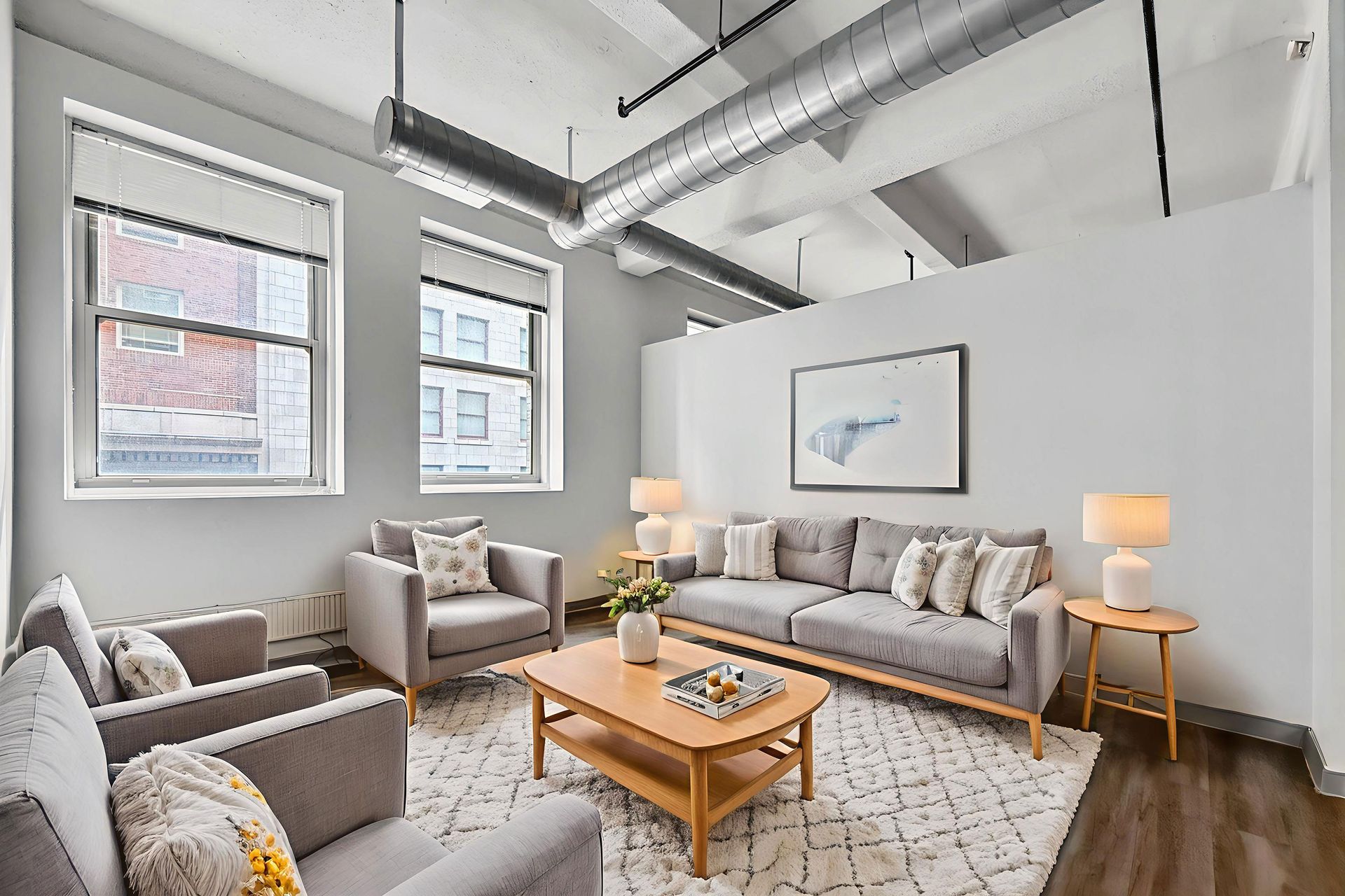 A modern living room with gray furniture, a wooden coffee table, a patterned rug, and industrial ceiling ductwork.