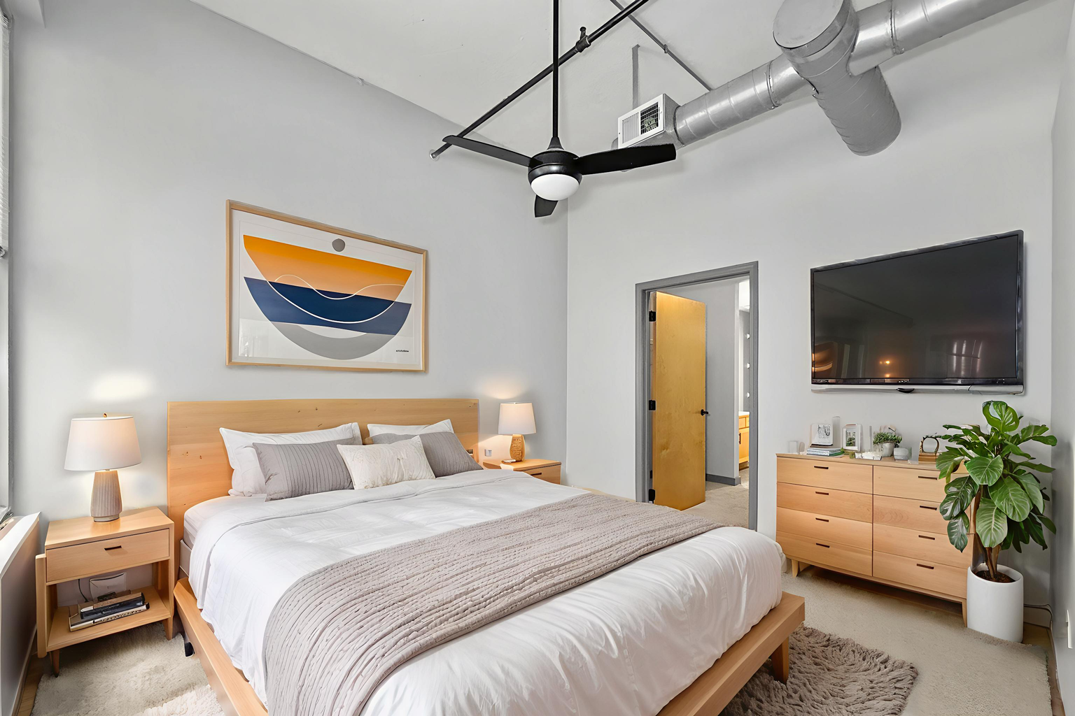 Modern bedroom with a light wood bed, matching dresser, large abstract wall art, and ceiling fan in an industrial space.
