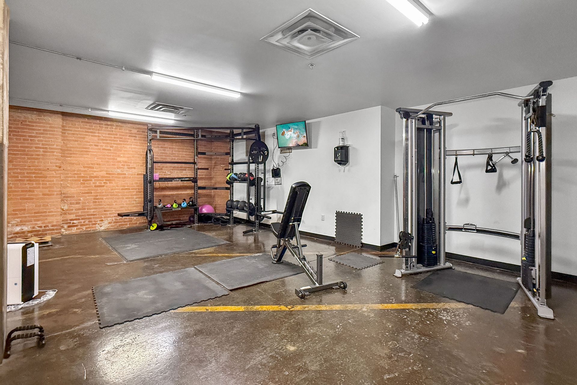 A modern gym with a brick wall, weight racks, a functional trainer machine, a bench, and gray rubber floor mats.