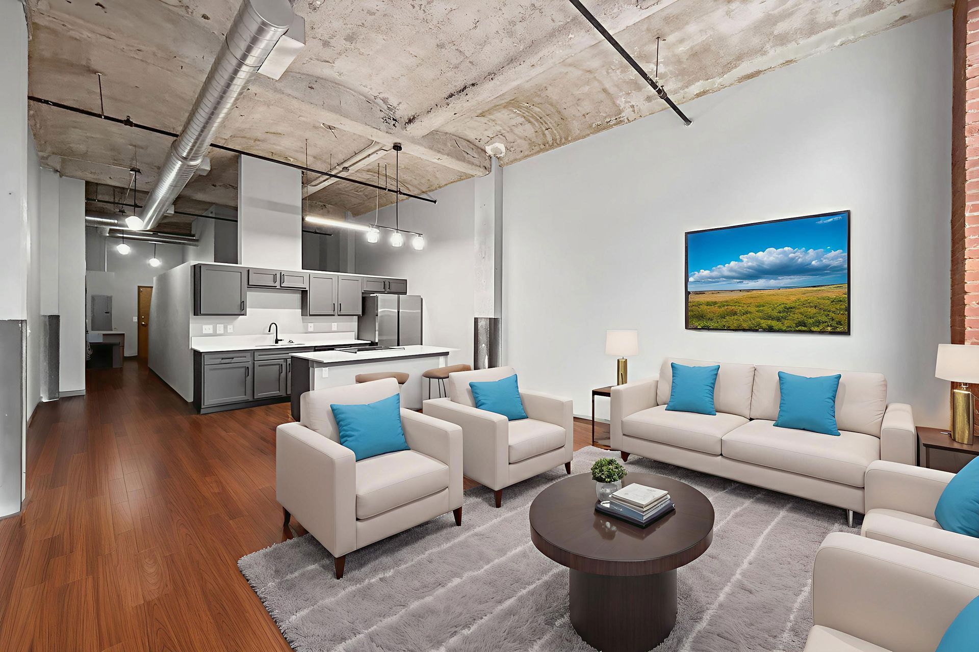 Open-concept living room with white sofas and blue pillows, a circular coffee table, and a kitchen area on concrete floors.