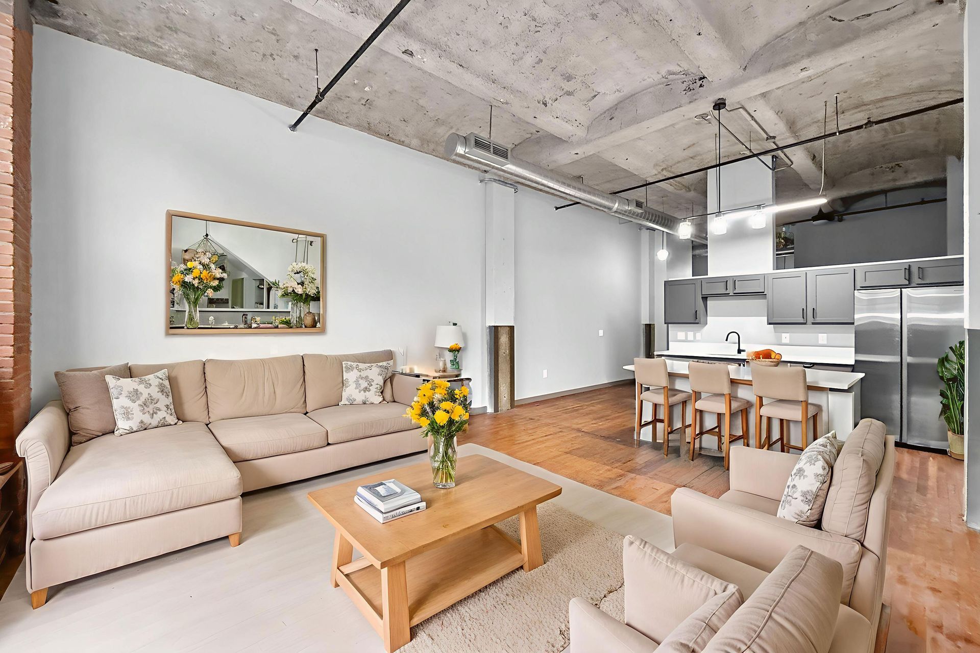 Open-plan living room with a beige sectional sofa, a wooden coffee table, and a kitchen with stainless steel appliances.
