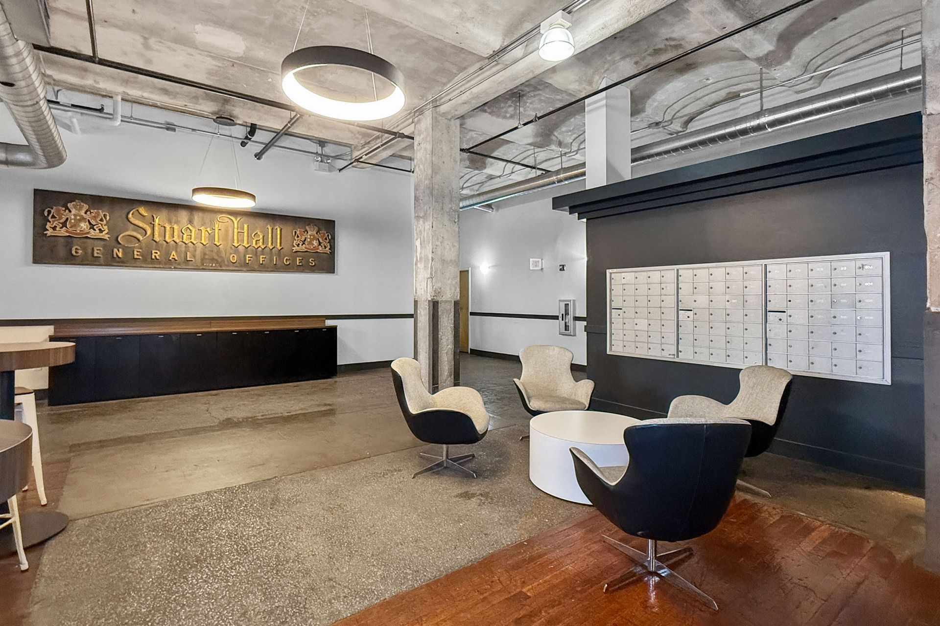 A modern lobby with a reception desk, lounge seating, and a mail center under industrial-style ceiling lights.
