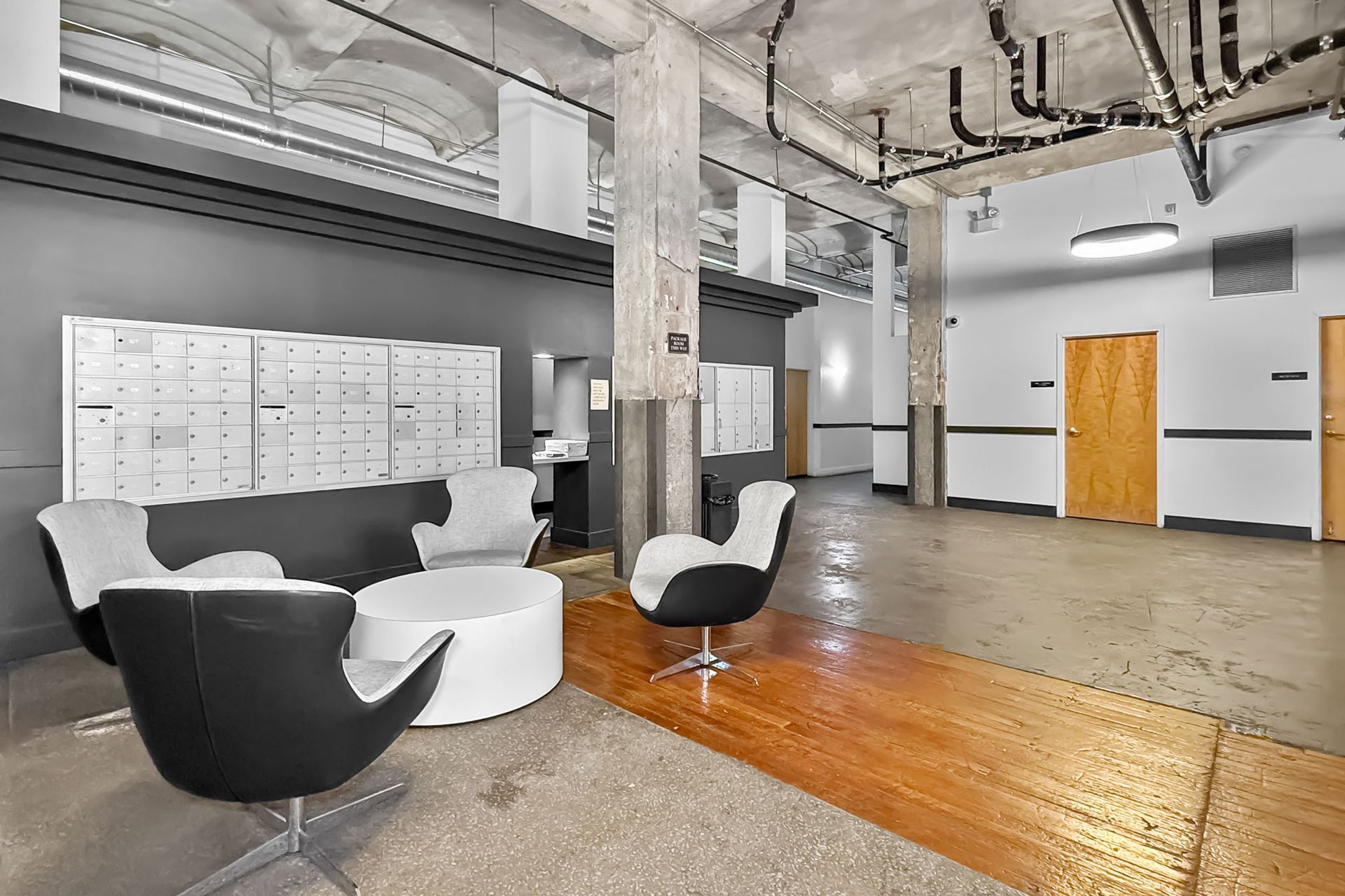 A modern, industrial lobby with white egg chairs, a circular white table, mailboxes, and exposed concrete pillars.