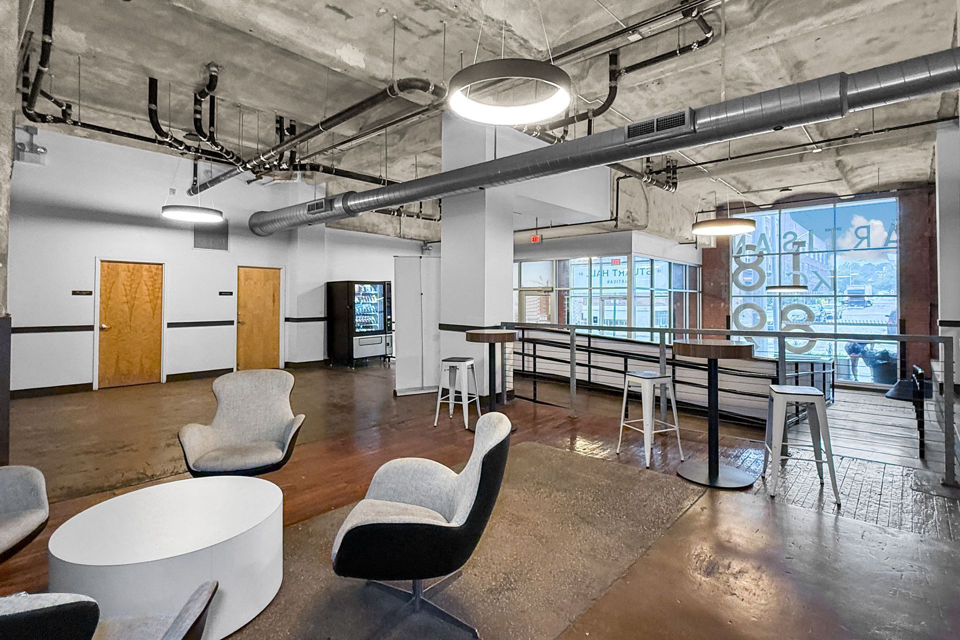 Modern office lounge with grey chairs, a white coffee table, exposed concrete ceiling, and large windows.