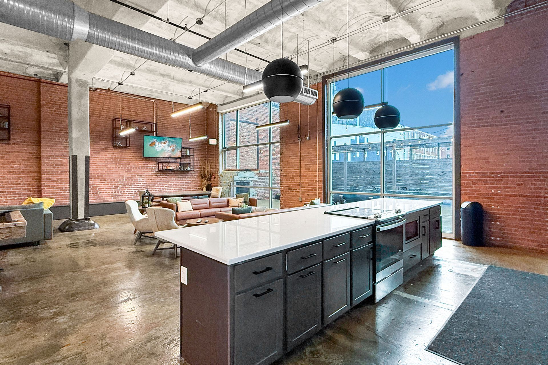 A modern industrial loft features a kitchen island, brick walls, concrete floors, and large windows with blue sky.