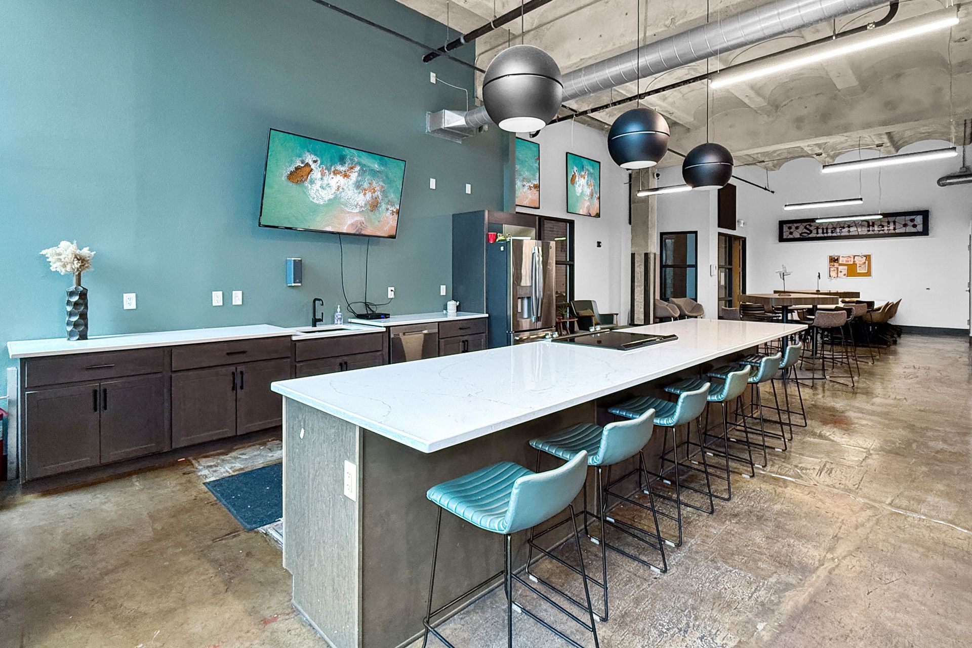 A modern office breakroom with a large kitchen island, teal chairs, dark cabinetry, and exposed industrial ceiling pipes.