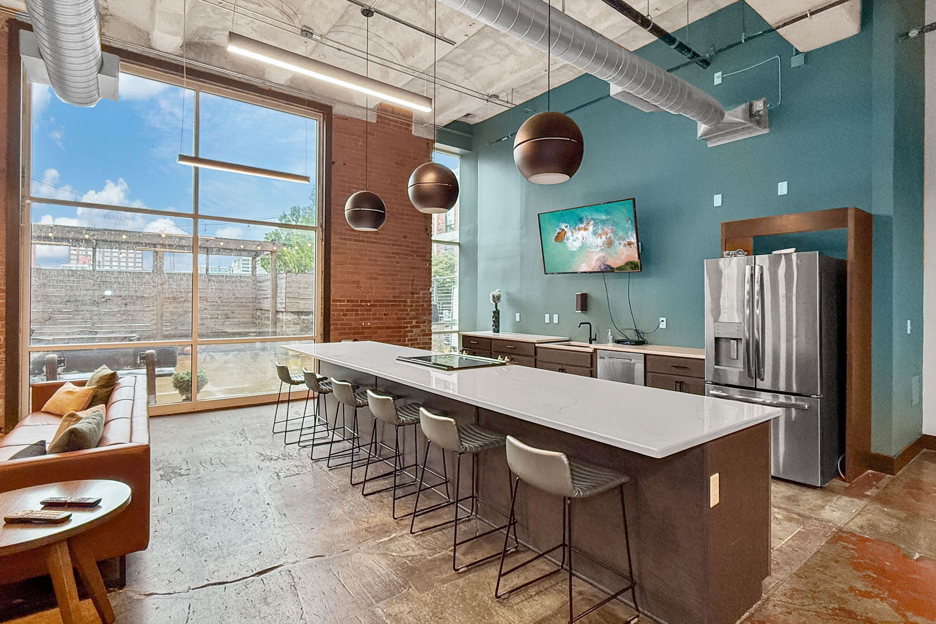 Modern lounge with a long white kitchen island, bar stools, stainless steel refrigerator, and large windows.