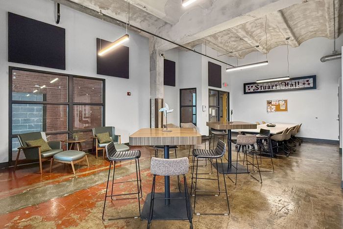 Modern office breakroom with high-top tables, bar stools, lounge chairs, and a long conference table on concrete floors.
