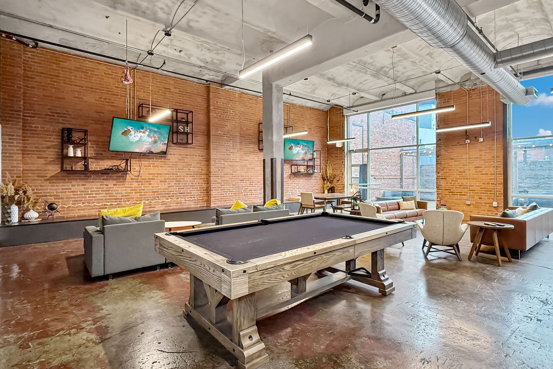 A spacious, modern lounge with exposed brick walls, a pool table, and contemporary seating under high industrial ceilings.