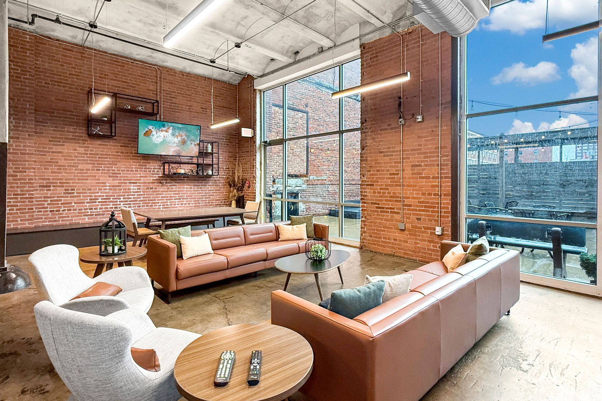A modern lounge with exposed brick walls, two leather sofas, white armchairs, and floor-to-ceiling windows.