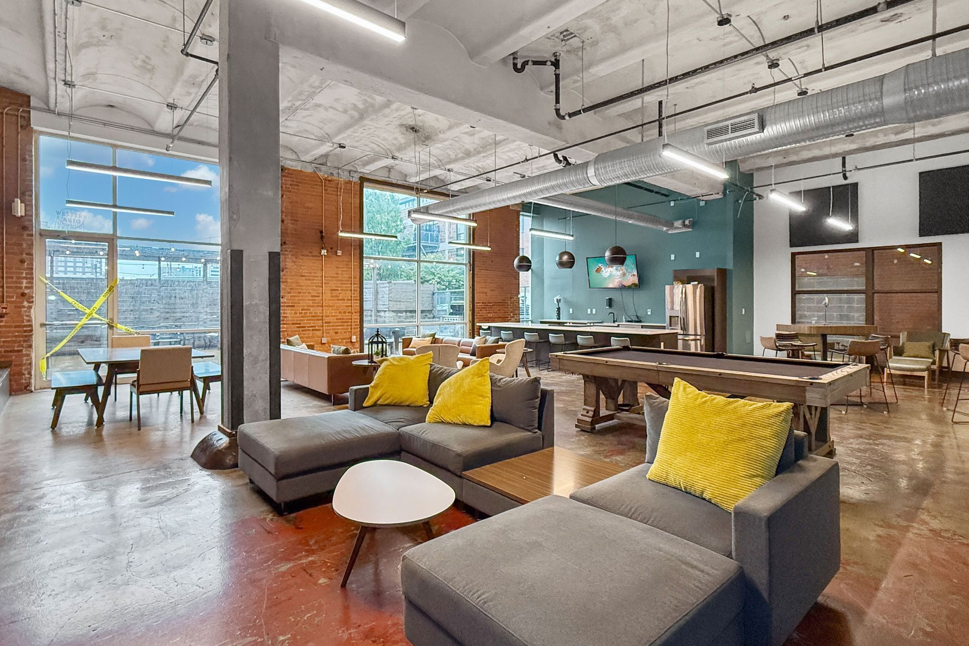 A modern, open-concept lounge with gray sectionals, bright yellow pillows, a pool table, and high-top seating.