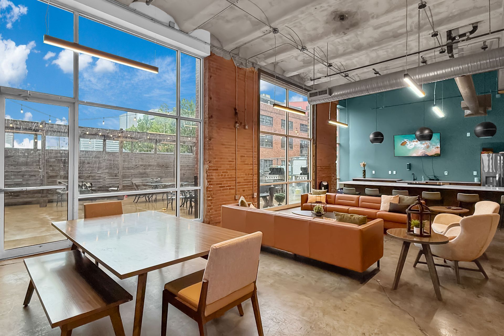 Modern industrial-style lounge with brick walls, floor-to-ceiling windows, leather couches, and a dining table.