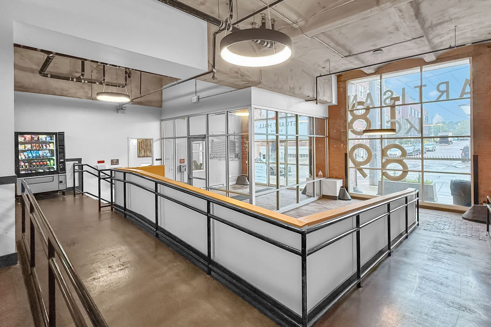 A modern interior space with a ramp leading to a glass-enclosed office, a vending machine, and large windows.