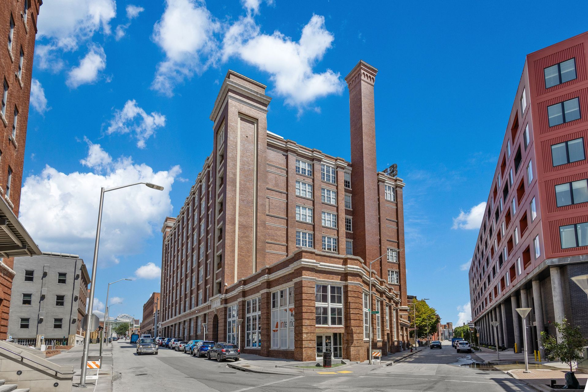 A street-level view of a tall, historic red-brick building with a prominent smokestack next to a modern red apartment block.