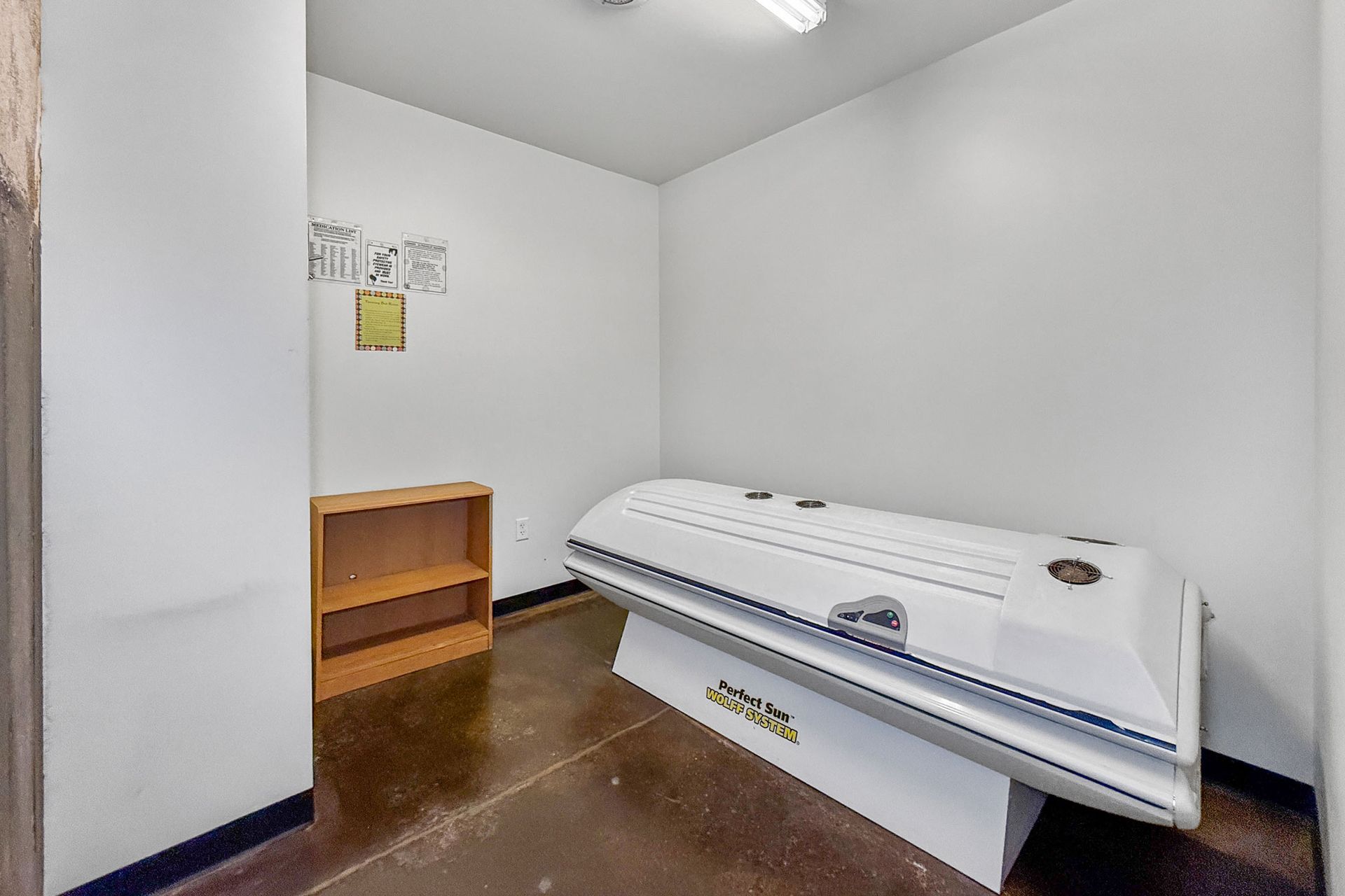 A white tanning bed sits in a corner of a sparse, white-walled room with a small wooden shelf and brown concrete flooring.