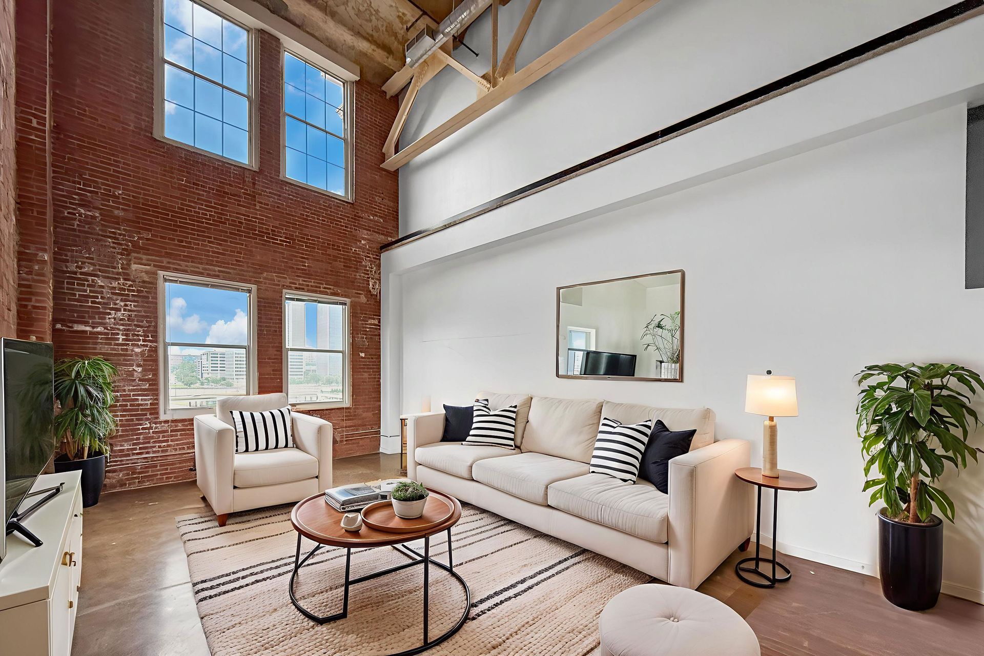 Living room with tall ceilings, exposed brick walls, a beige sofa, armchair, coffee table, and large windows.