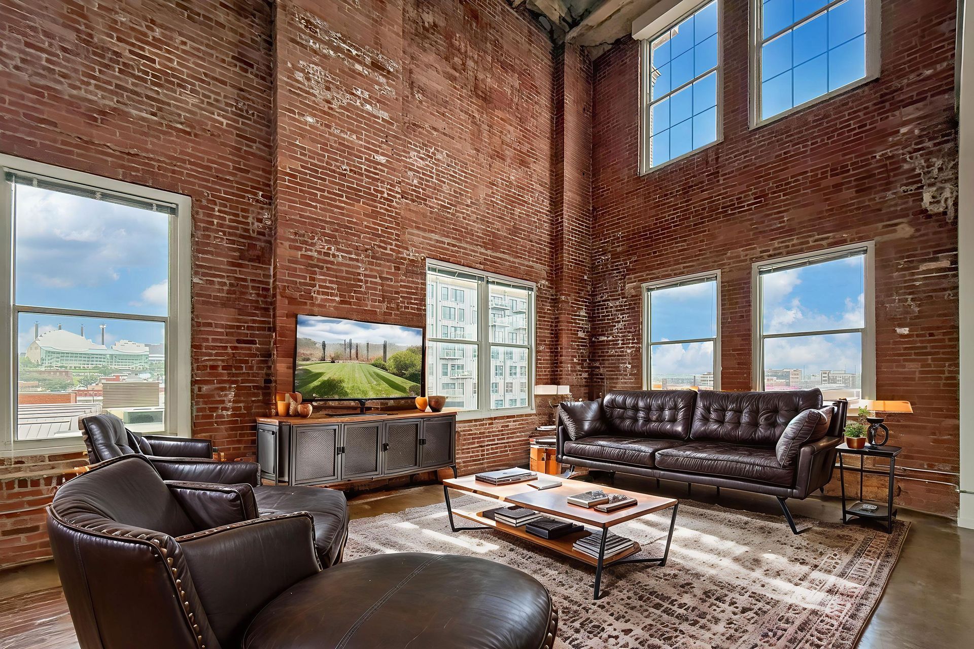 A loft living room with exposed red brick walls, high ceilings, large windows, dark leather furniture, and a rug.