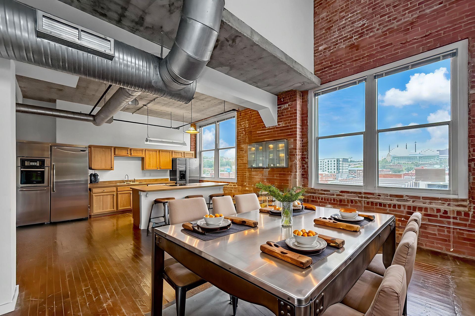 A modern open-plan dining and kitchen area with a metal table, industrial ceiling vents, and exposed brick walls.